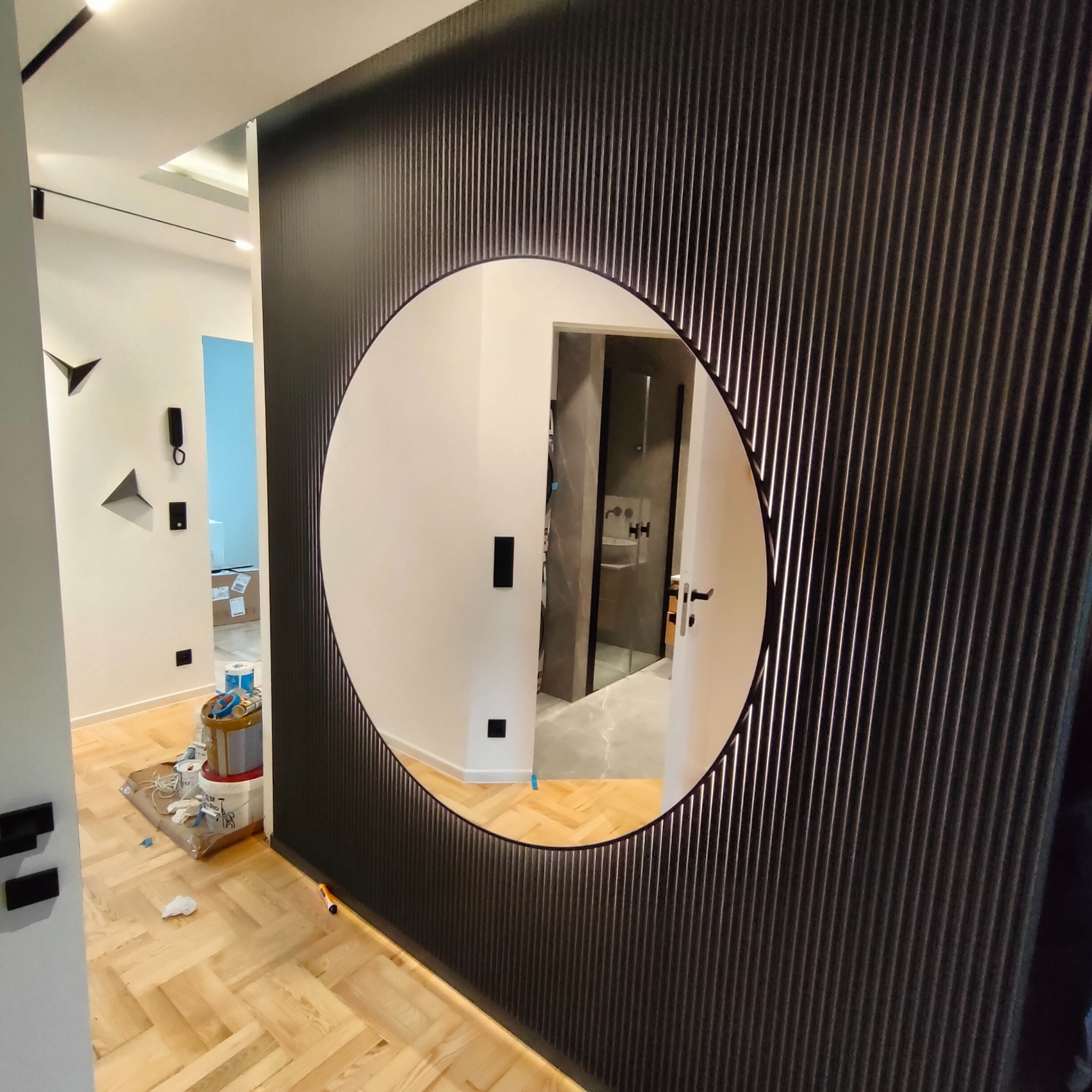 Nowoczesne wnętrze z ciemną ścianą z pionowymi żłobieniami i okrągłym lustrem, w którym odbija się łazienka; na podłodze widoczne resztki po malowaniu i narzędzia malarskie.