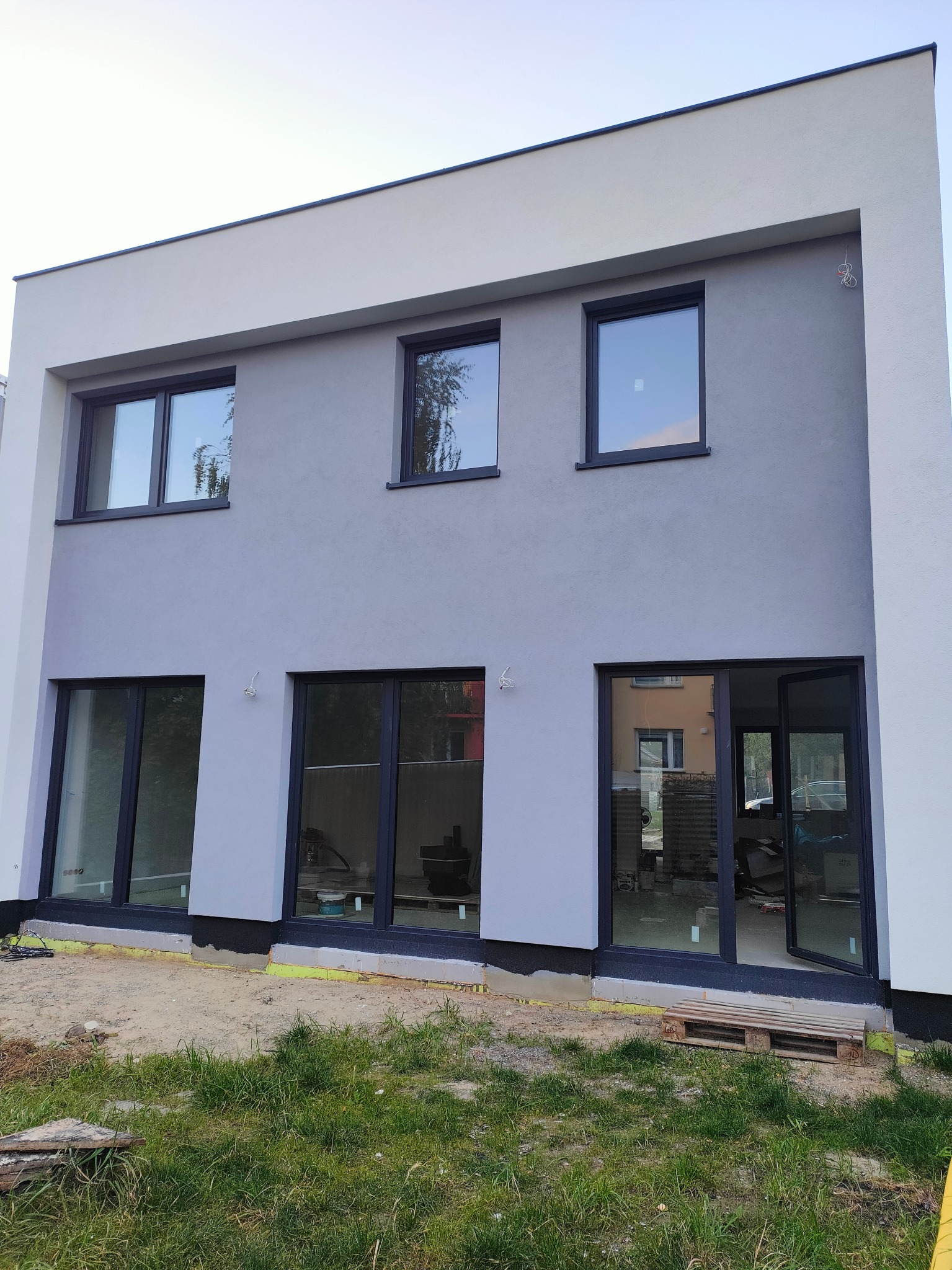 Dwukondygnacyjny dom jednorodzinny w Lublinie z szarą elewacją i czarną stolarką okienną, widoczne ślady prac wykończeniowych wokół budynku.