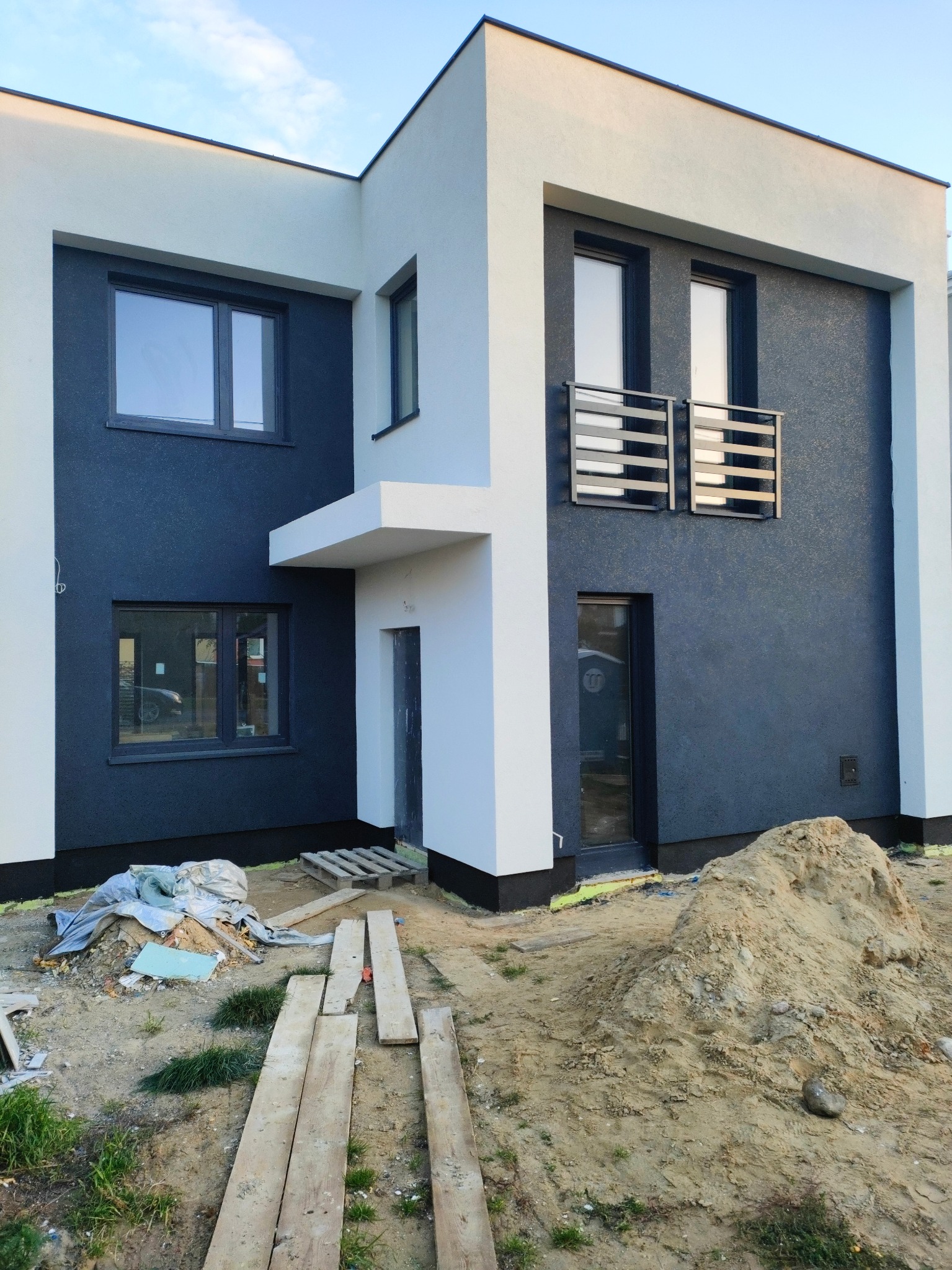 Dwukondygnacyjny dom z nowoczesną elewacją w kolorach bieli i grafitu, widoczne okna z grafitowymi ramami, mały balkon z metalową balustradą oraz teren wokół domu w trakcie prac wykończeniowych,...