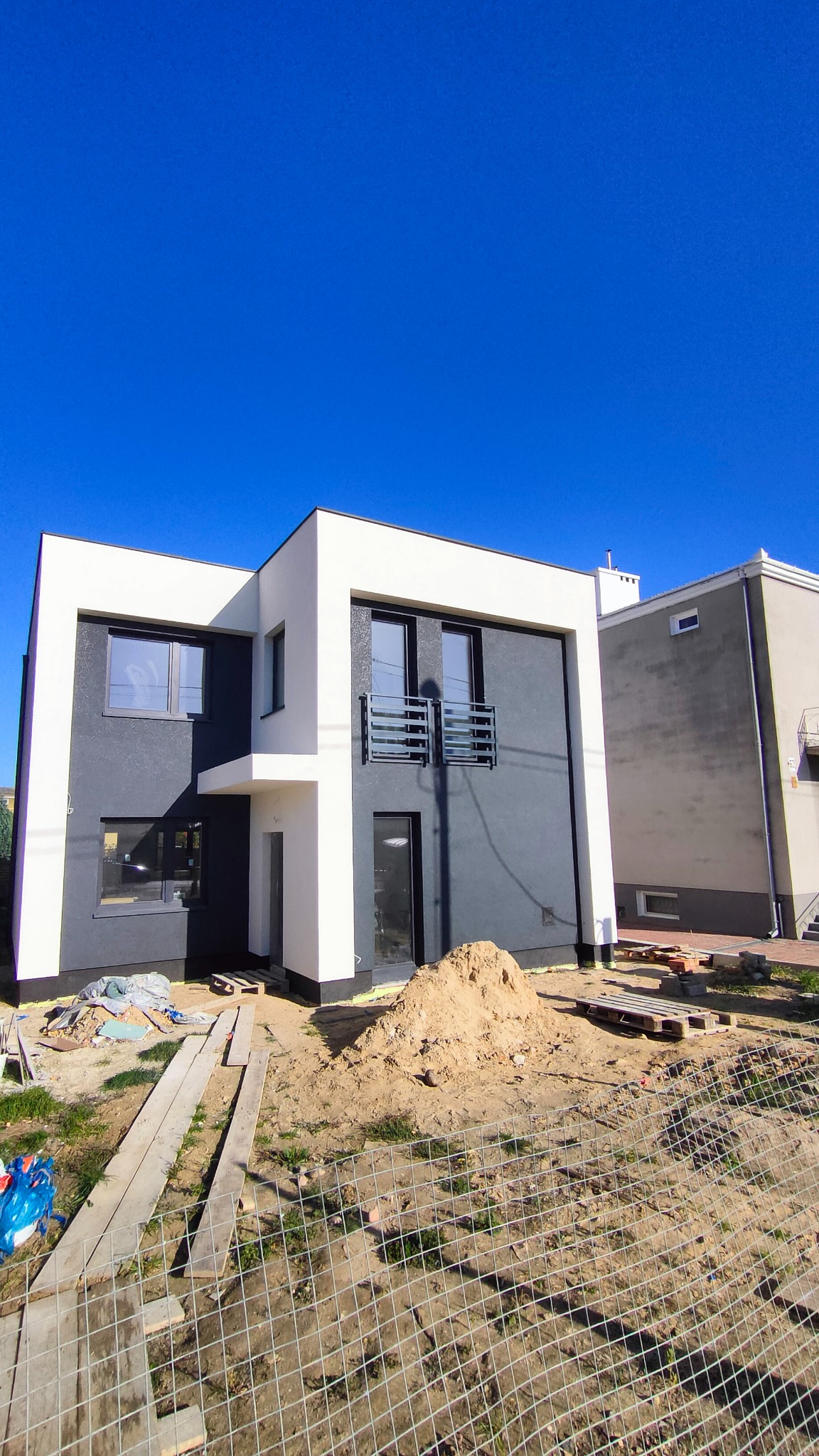 Nowoczesny dom jednorodzinny z elewacją w kolorach bieli i grafitu, widok z dołu, teren wokół w trakcie prac wykończeniowych, widoczna sterta piasku i tymczasowe ogrodzenie.