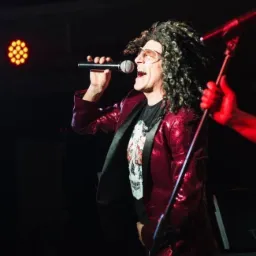 Wokalista w błyszczącej czerwonej marynarce i kręconej peruce śpiewa do mikrofonu na scenie oświetlonej czerwonym światłem.