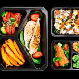 Zestaw posiłków w czarnych pojemnikach: kurczak z przyprawami i truskawkami na szpinaku, bataty, sałatka z awokado i pomidorów, łosoś z brokułami i komosą ryżową, kasza z warzywami i tofu.