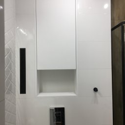 Tabre Sp. z o.o. - Biała ściana w łazience z ukrytą szafką z wnęką, chromowanym przyciskiem spłuczki i czarnymi kratkami wentylacyjnymi.