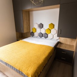 Tabre Sp. z o.o. - Nowoczesna sypialnia z łóżkiem, szafkami nocnymi i szafą w zabudowie w odcieniach szarości i drewna, z żółtą narzutą i heksagonalnym zagłówkiem.