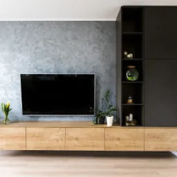 Nowoczesny salon z wiszącą szafką RTV z drewnianym frontem, grafitową ścianą z efektem dekoracyjnym, czarnymi regałami i roślinami doniczkowymi.