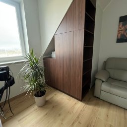 Tabre Sp. z o.o. - Drewniana szafa wnękowa z półkami, dopasowana do skosu dachu, obok okna i sofy, z rośliną doniczkową na pierwszym planie.