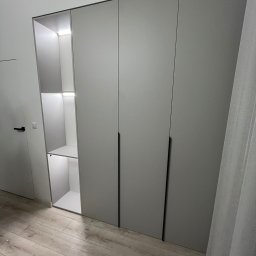 Tabre Sp. z o.o. - Szara szafa wnękowa z oświetleniem LED wewnątrz, minimalistyczne czarne uchwyty, drewniana podłoga, widok z boku, zasłona w tle.