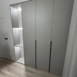 Szara szafa wnękowa z oświetleniem LED wewnątrz, minimalistyczne czarne uchwyty, drewniana podłoga, widok z boku, zasłona w tle.