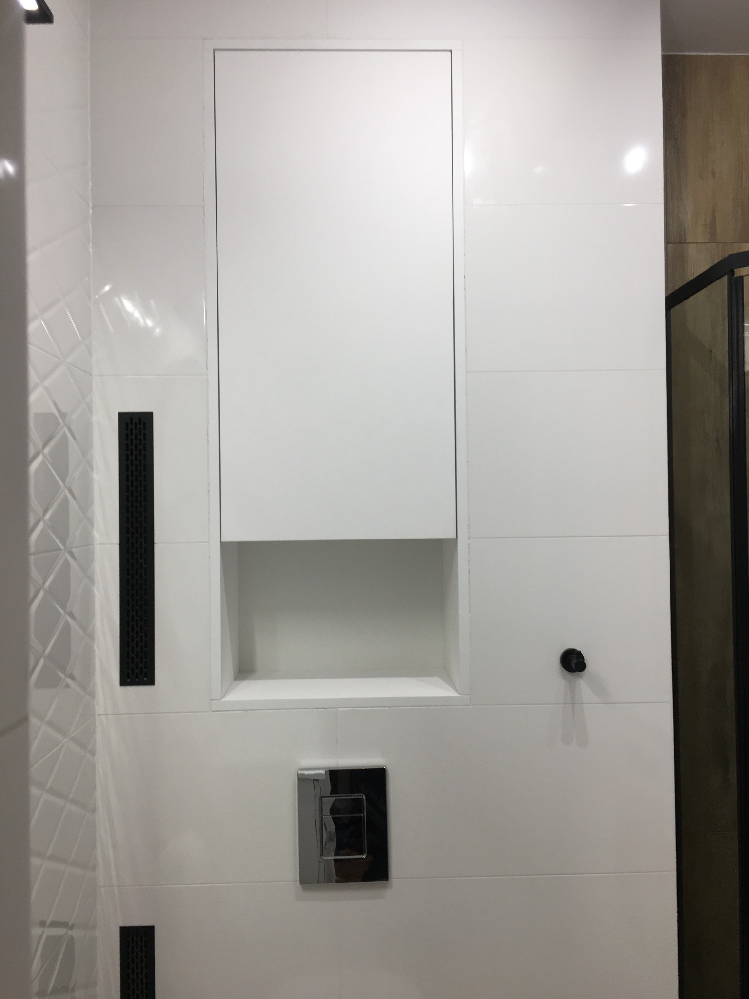 Biała ściana w łazience z ukrytą szafką z wnęką, chromowanym przyciskiem spłuczki i czarnymi kratkami wentylacyjnymi.