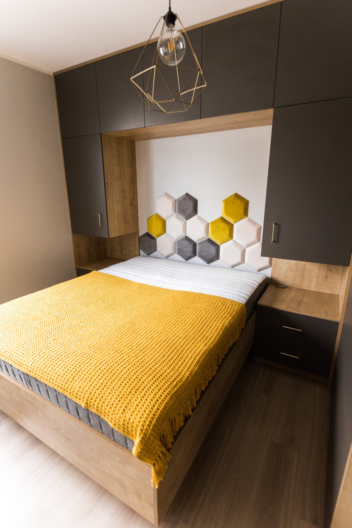 Nowoczesna sypialnia z łóżkiem, szafkami nocnymi i szafą w zabudowie w odcieniach szarości i drewna, z żółtą narzutą i heksagonalnym zagłówkiem.