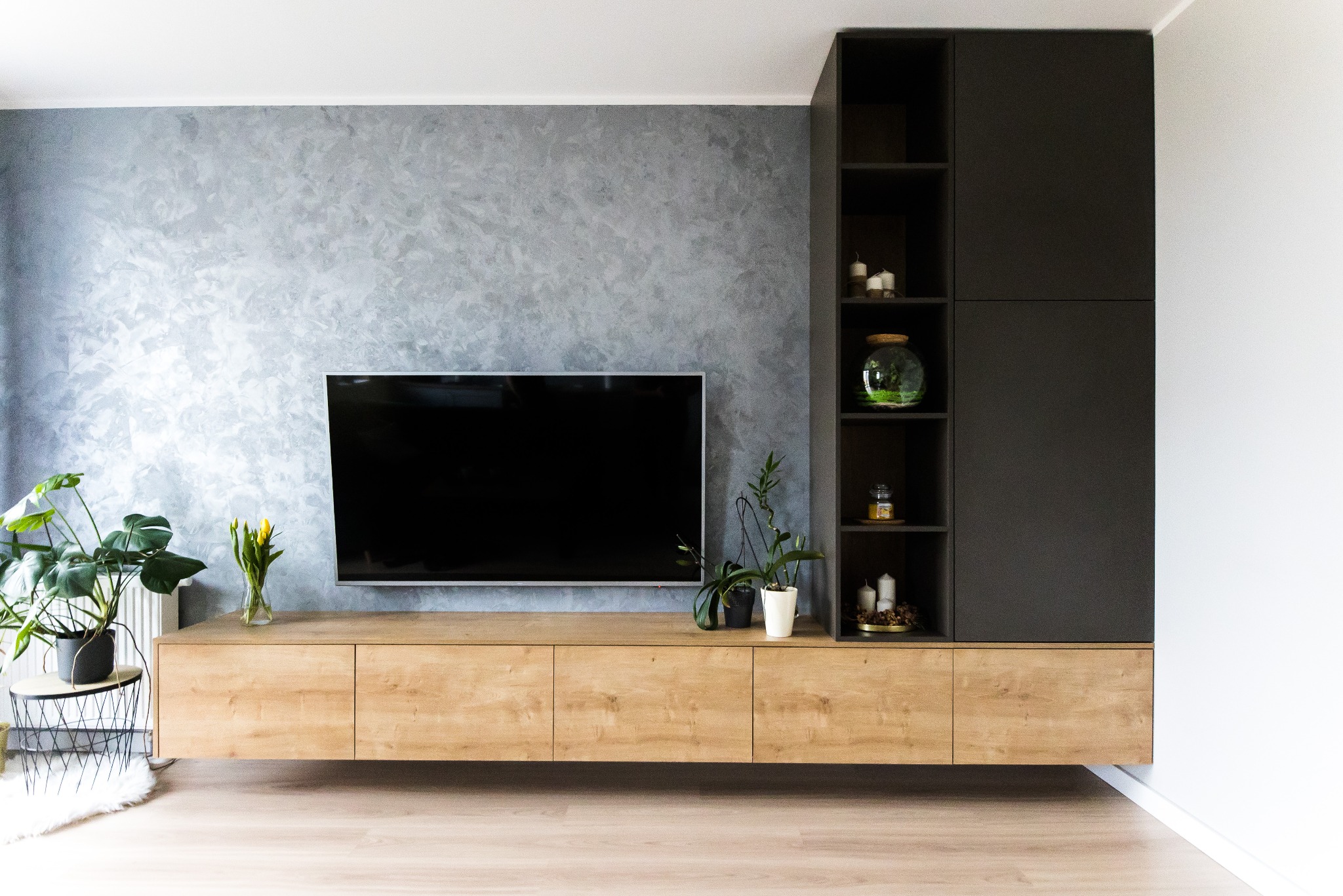 Nowoczesny salon z wiszącą szafką RTV z drewnianym frontem, grafitową ścianą z efektem dekoracyjnym, czarnymi regałami i roślinami doniczkowymi.