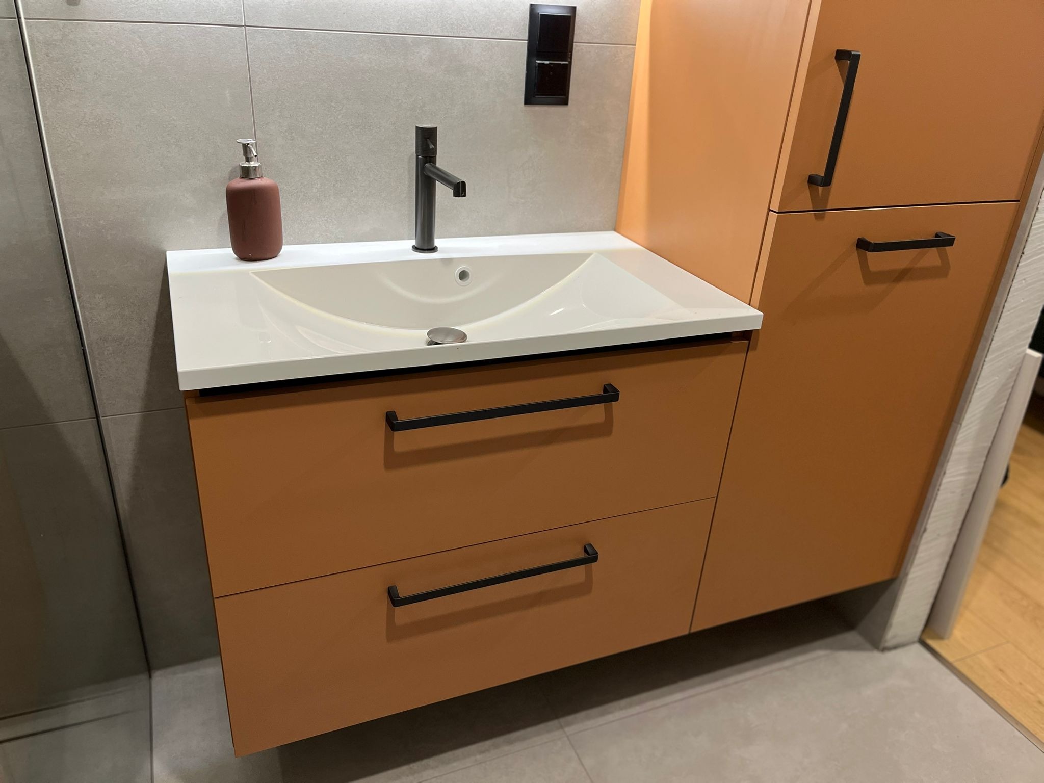 Nowoczesna łazienka z pomarańczową szafką pod umywalkę z szufladami i wysoką szafką boczną, czarna bateria umywalkowa i podłużne uchwyty, szare płytki na ścianie i podłodze.