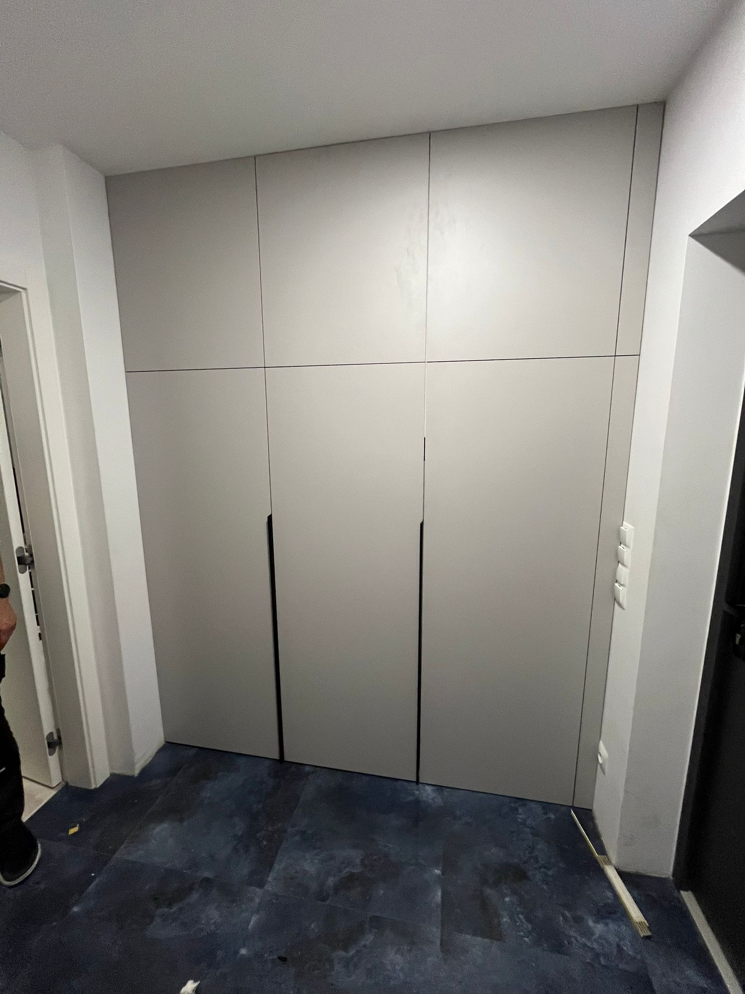 Szafa wnękowa z szarymi frontami, czarnymi uchwytami, zamontowana od podłogi do sufitu, widoczne listwy przypodłogowe i fragment podłogi w odcieniach granatu.