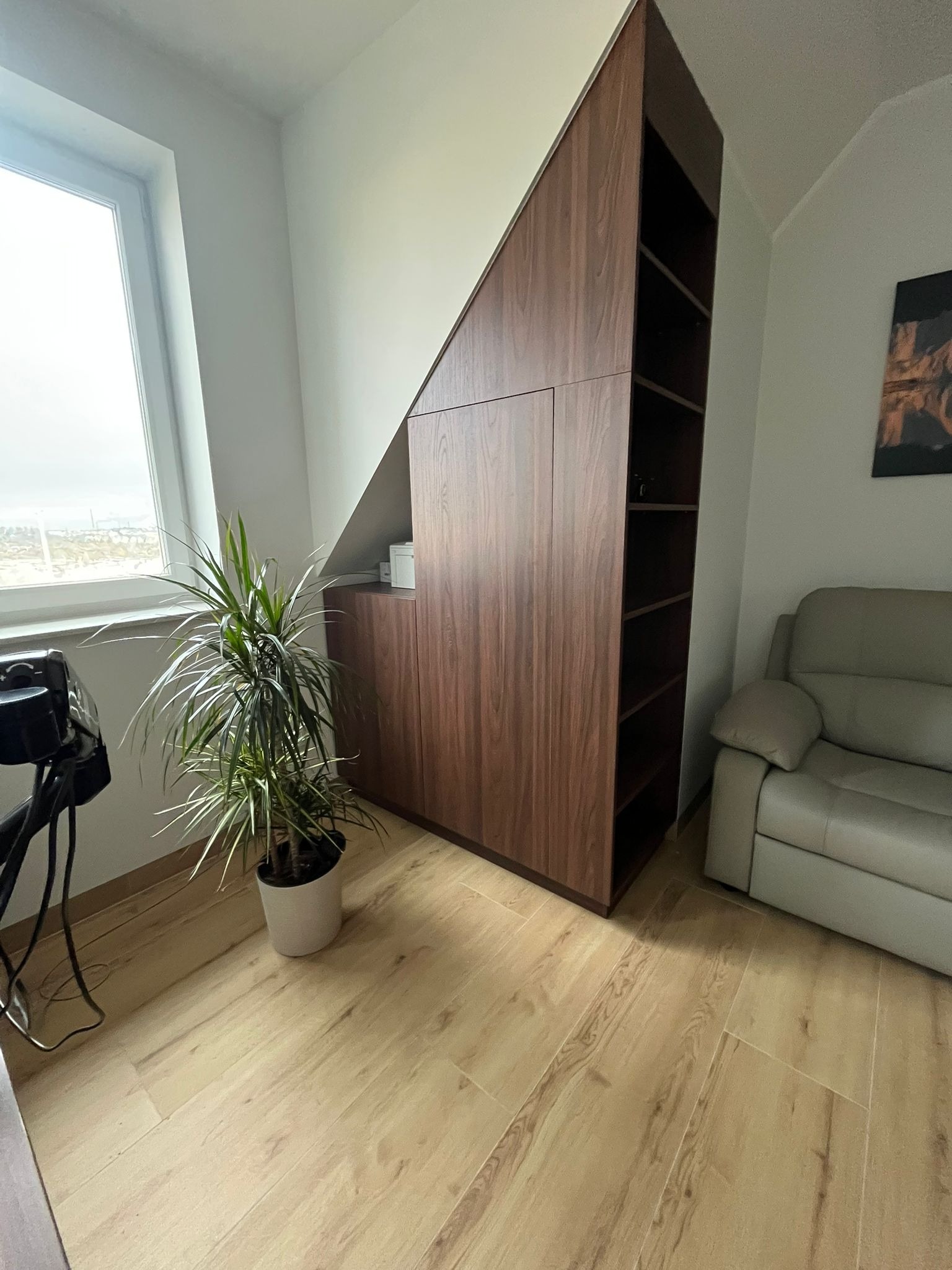 Drewniana szafa wnękowa z półkami, dopasowana do skosu dachu, obok okna i sofy, z rośliną doniczkową na pierwszym planie.