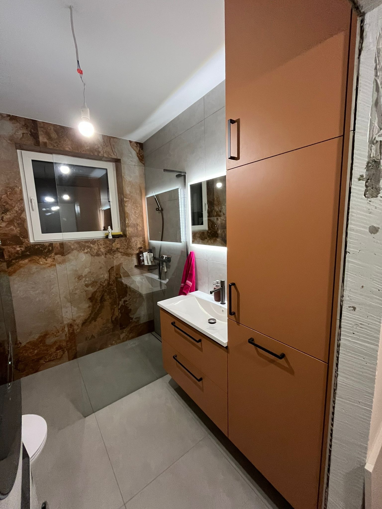 Nowoczesna łazienka w trakcie wykańczania, z brązową szafką pod umywalką i lustrem z podświetleniem LED, widoczny fragment ściany z niedokończonym tynkiem, oraz surowa żarówka zwisająca z sufitu.