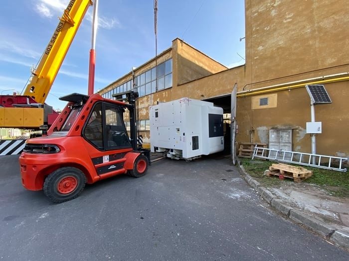 Rozładunek frezarki CNC o wadze 21 ton przy użyciu wózka widłowego Linde i dźwigu, z ustawieniem maszyny w nowej lokalizacji, widoczne palety i drabina oparta o budynek.