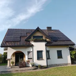 Biały dom jednorodzinny z ciemną dachówką, na której zamontowane są panele słoneczne; widoczne również rolety zewnętrzne w oknach, taras z meblami ogrodowymi i zadbany trawnik.