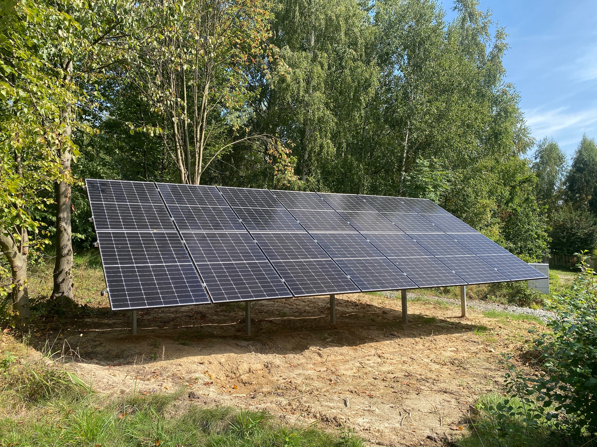Instalacja paneli słonecznych na metalowej konstrukcji, otoczona drzewami i przygotowanym terenem z piaskiem.