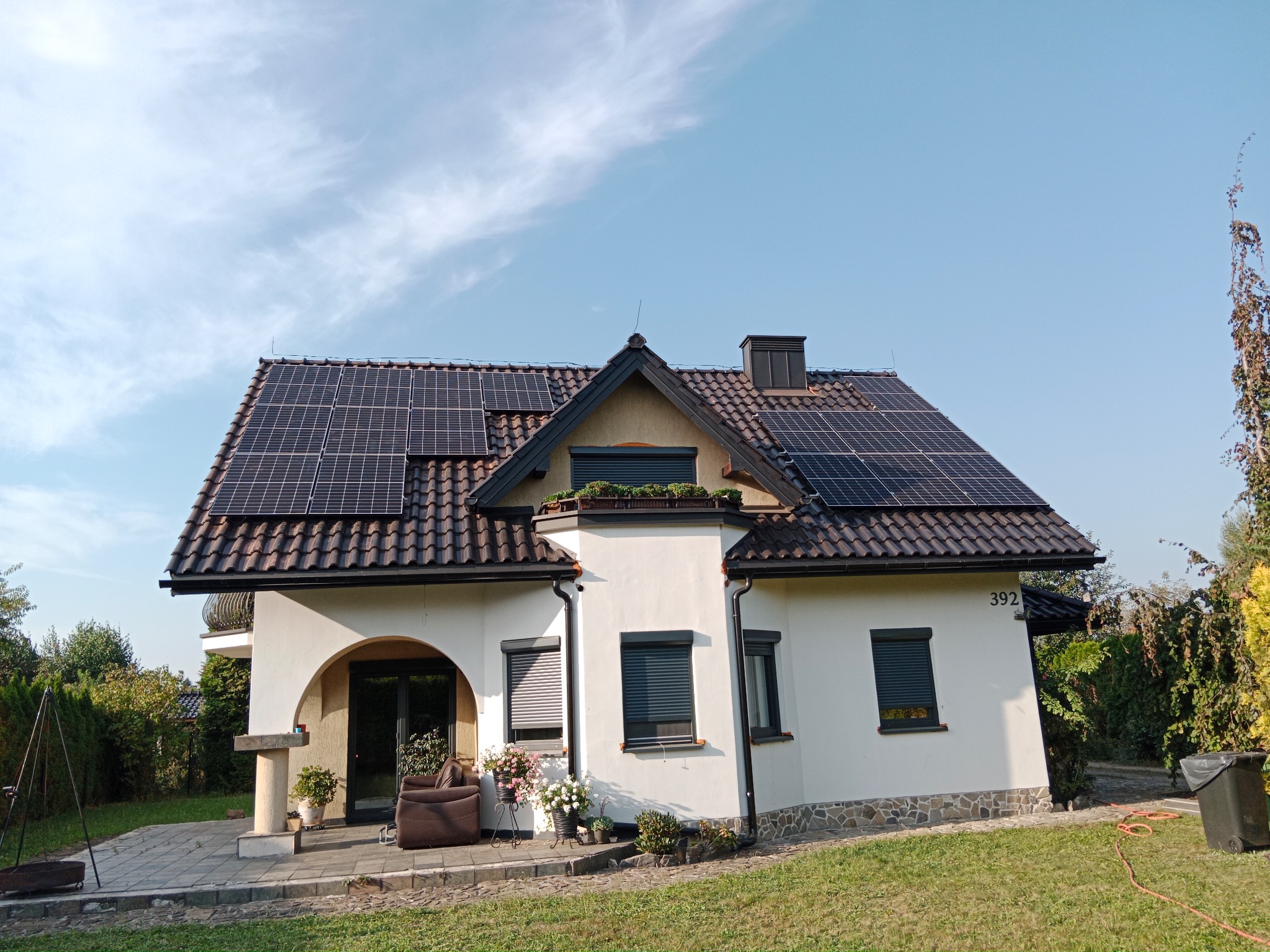 Biały dom jednorodzinny z ciemną dachówką, na której zamontowane są panele słoneczne; widoczne również rolety zewnętrzne w oknach, taras z meblami ogrodowymi i zadbany trawnik.