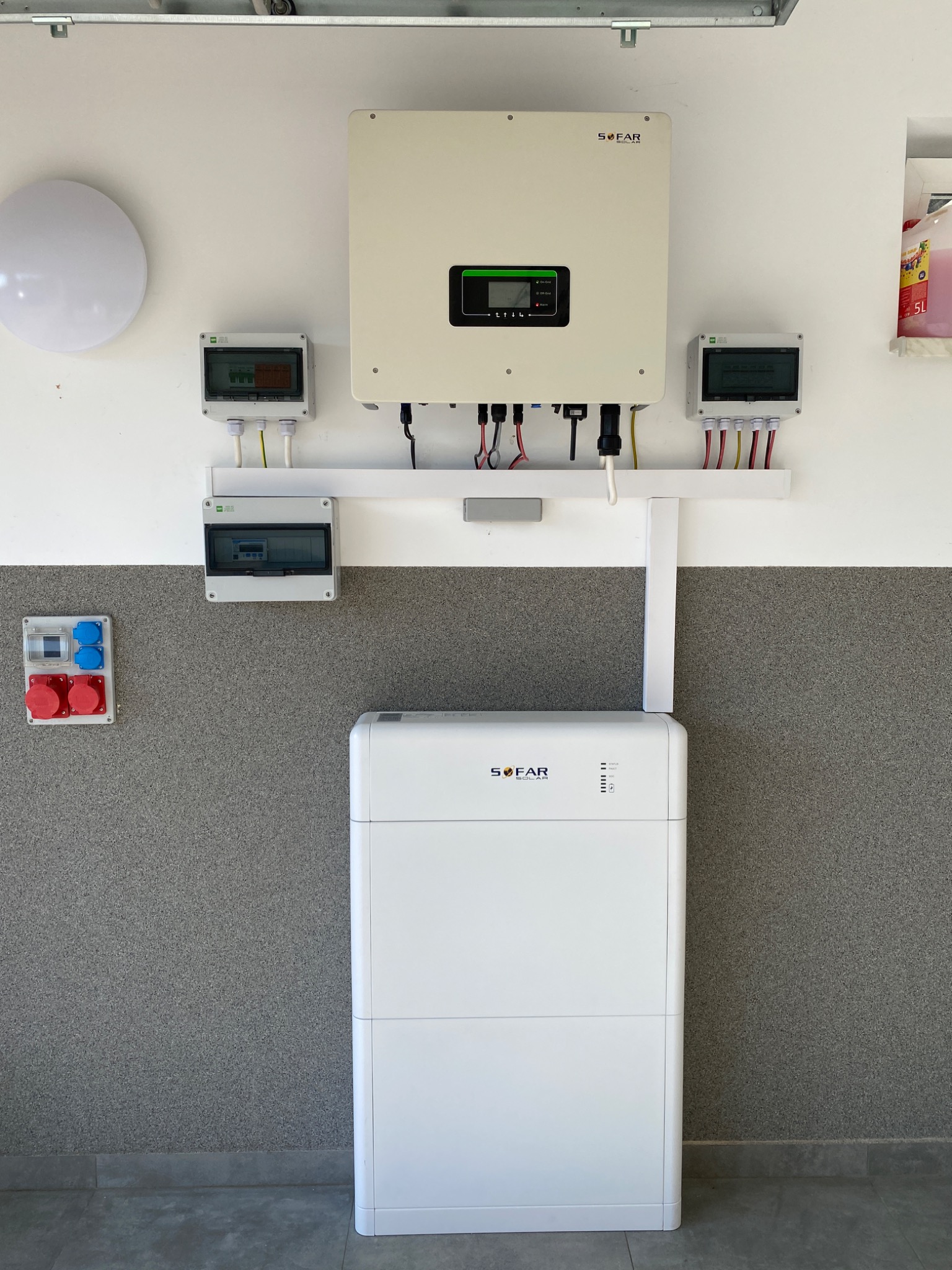 Biała bateria energii z falownikiem i skrzynkami elektrycznymi zamontowana na dwukolorowej ścianie w garażu.