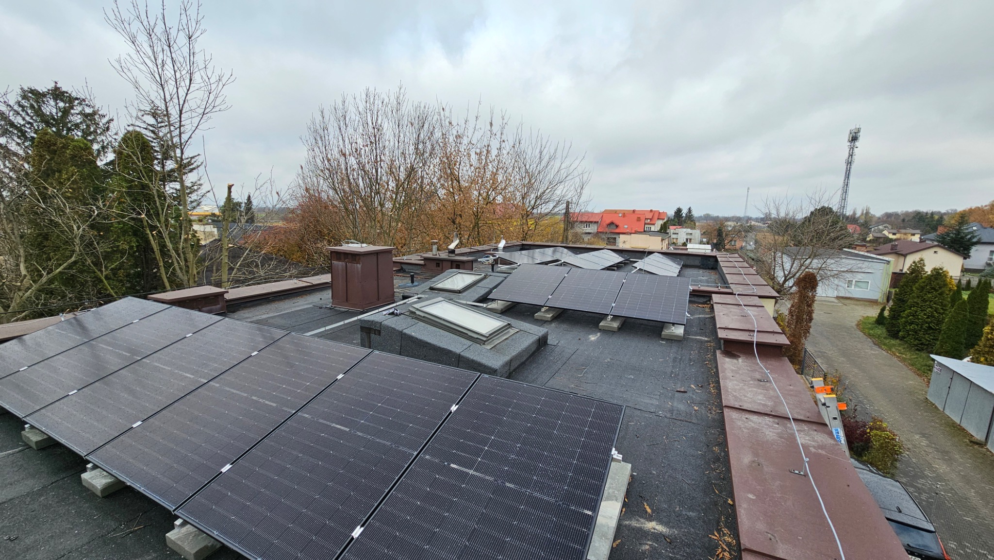 Instalacja paneli słonecznych na płaskim dachu z widocznymi świetlikami, kominami wentylacyjnymi, elementami mocującymi i fragmentem drabiny na tle zabudowy miejskiej w pochmurny dzień.