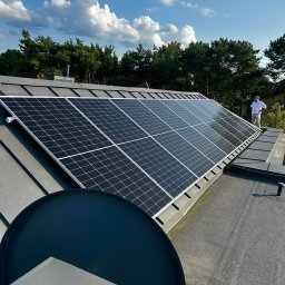 MCW Energia - Panel fotowoltaiczny na szarym dachu domu jednorodzinnego, widoczny pracownik w białej koszuli na tle błękitnego nieba z chmurami i zieleni drzew