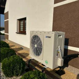 Zewnętrzna jednostka klimatyzacji marki York zamontowana na metalowym stelażu przy ścianie domu z brązowymi detalami, obok zadbane krzewy.