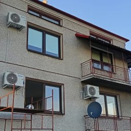 MCW Energia - Elewacja domu z zainstalowanymi klimatyzatorami Panasonic, widoczne okna z brązowymi ramami, balkon z metalową balustradą i rusztowanie przy ścianie.