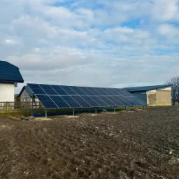 Instalacja fotowoltaiczna gruntowa z czarnymi panelami na metalowym stelażu, w tle budynki mieszkalne i pole orne pod pochmurnym niebem.
