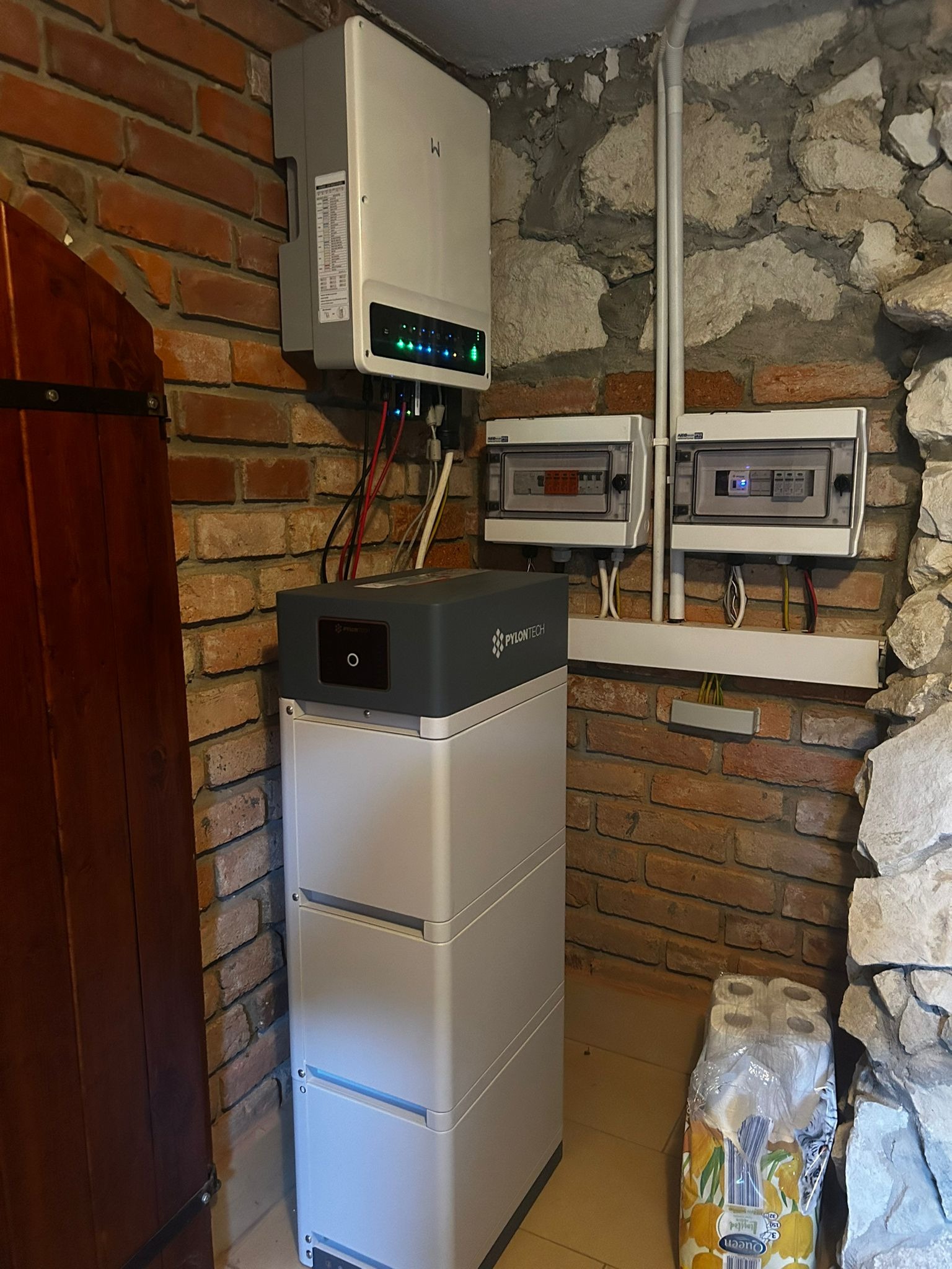 Instalacja z akumulatorem Pylontech, inwerterem z diodami sygnalizacyjnymi i rozdzielnicami elektrycznymi na tle ściany z cegły i kamienia