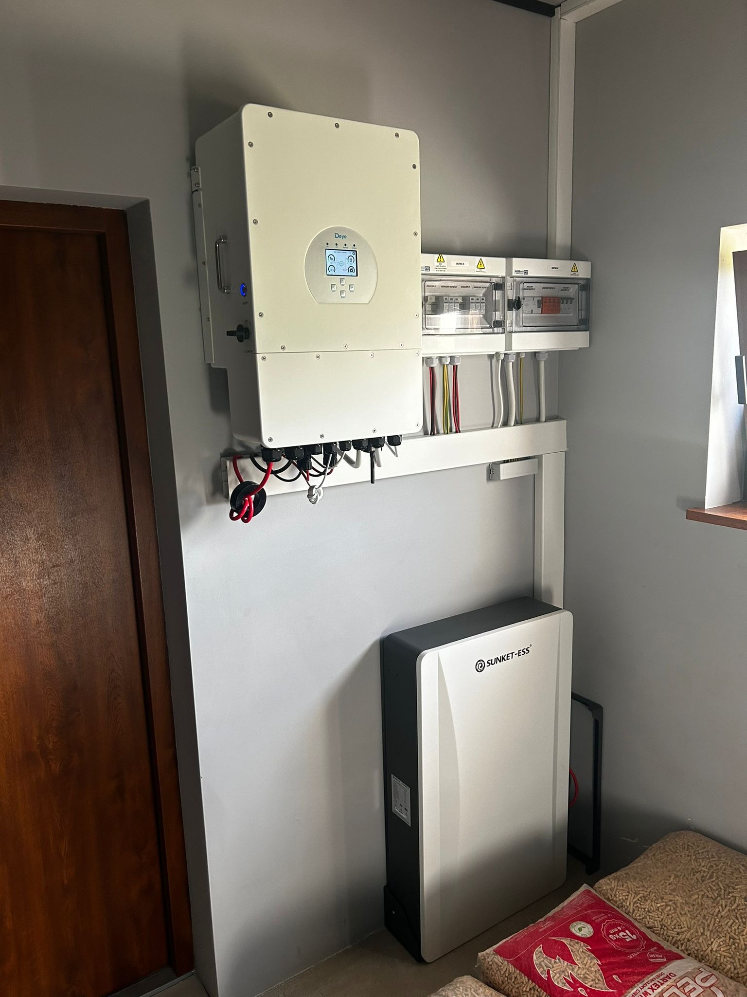 Biały inwerter z wyświetlaczem i skrzynka bezpiecznikowa zamontowane na szarej ścianie obok brązowych drzwi, poniżej akumulator SUNKET-ESS i worki z pelletem.