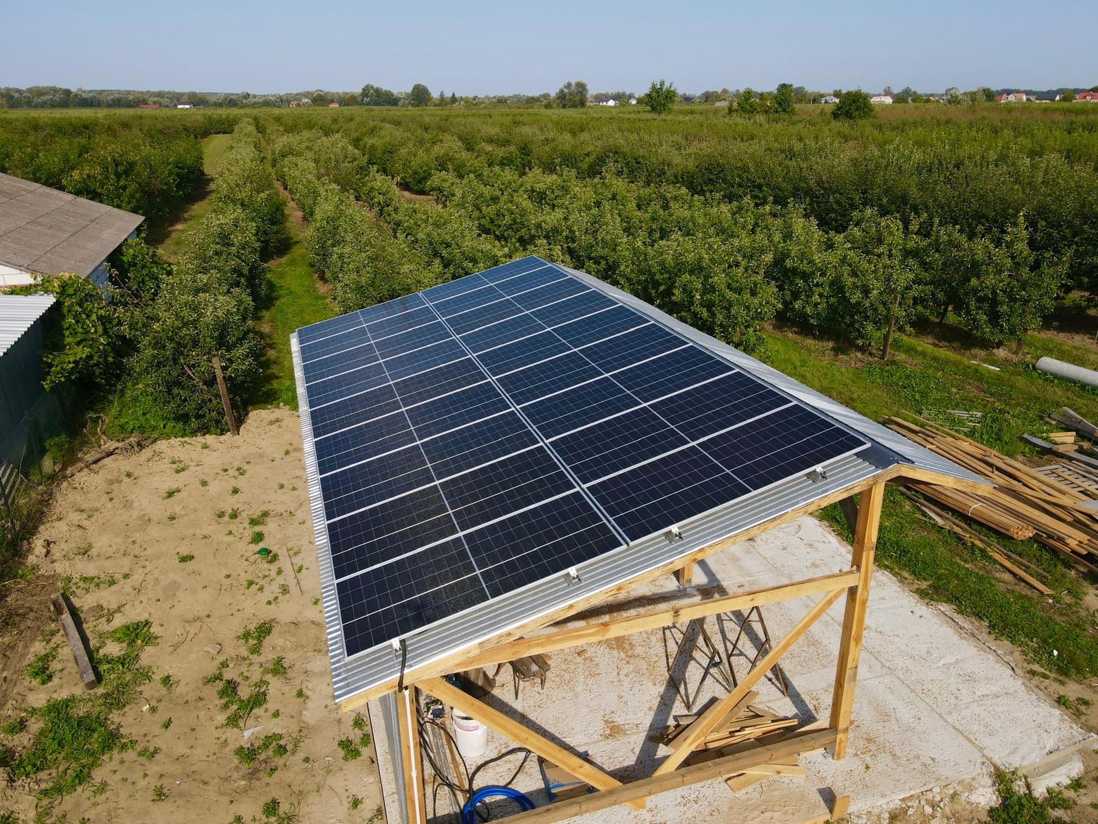 Instalacja fotowoltaiczna na drewnianej konstrukcji z blachy falistej, widok z góry na tle sadu owocowego.