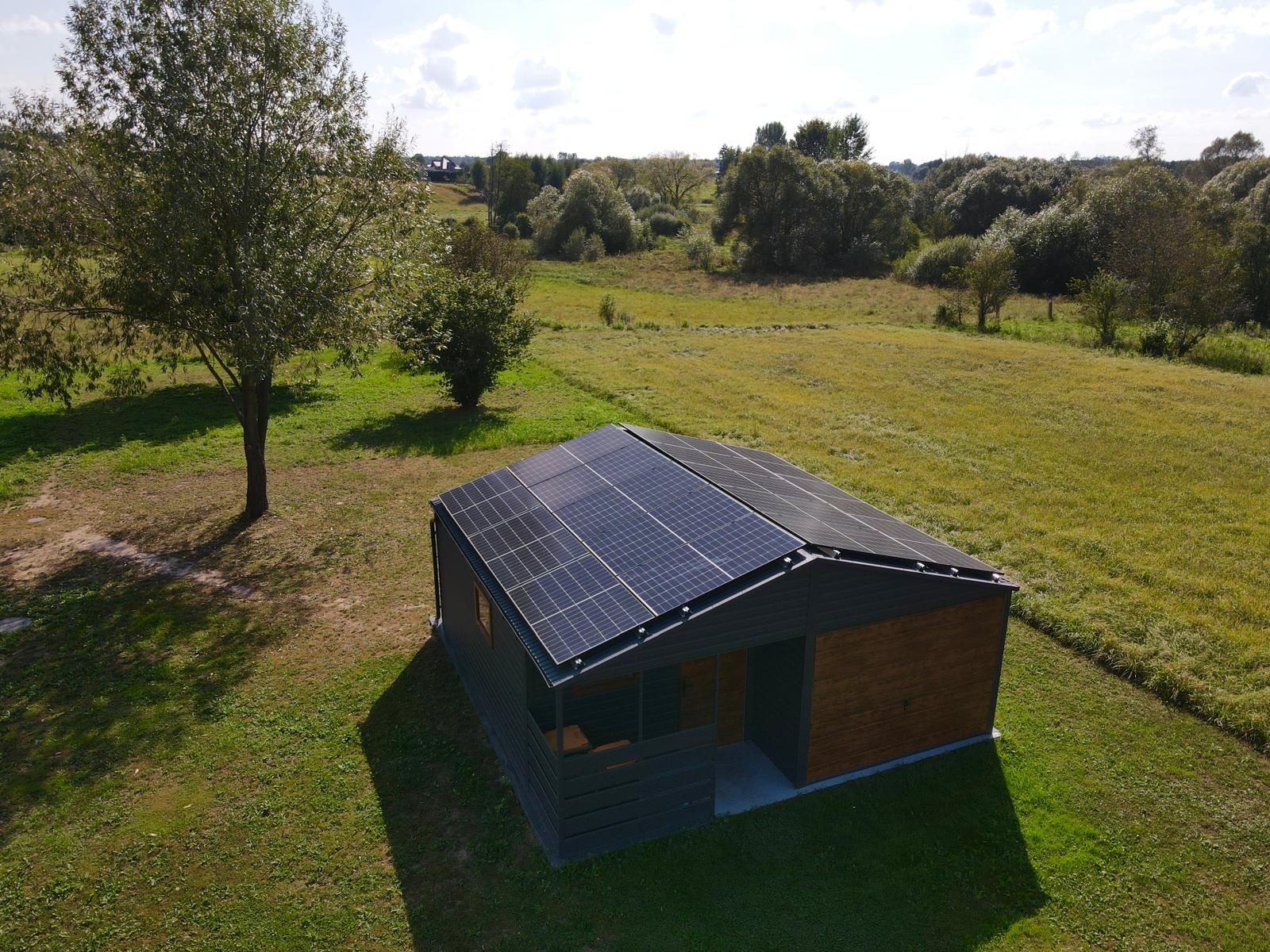 Niewielki, nowoczesny budynek z panelami fotowoltaicznymi na dachu, otoczony zielonym trawnikiem i drzewami, widok z lotu ptaka w słoneczny dzień.
