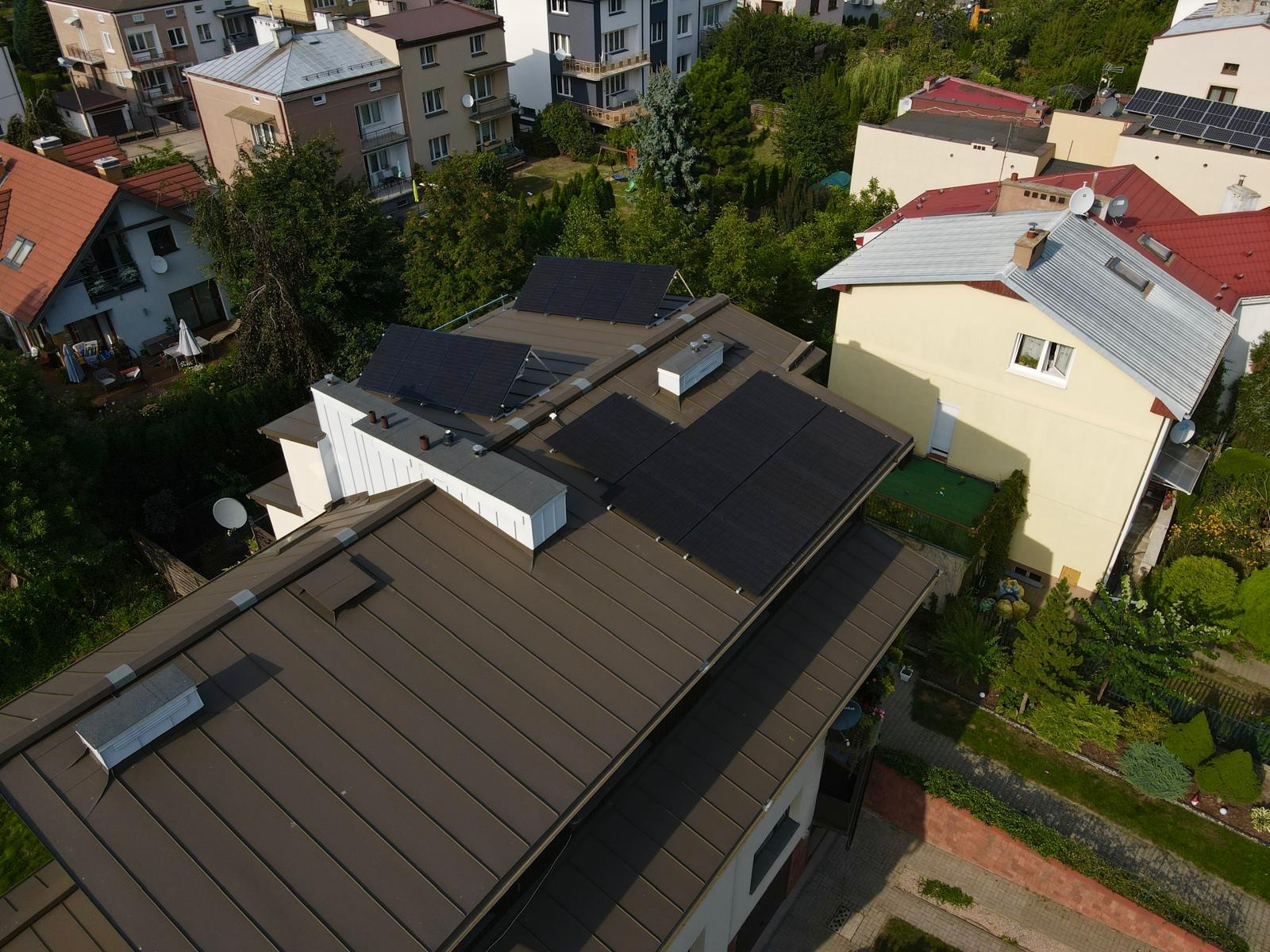 Widok z góry na brązowy dach z zainstalowanymi panelami fotowoltaicznymi na tle zabudowy jednorodzinnej i zieleni.