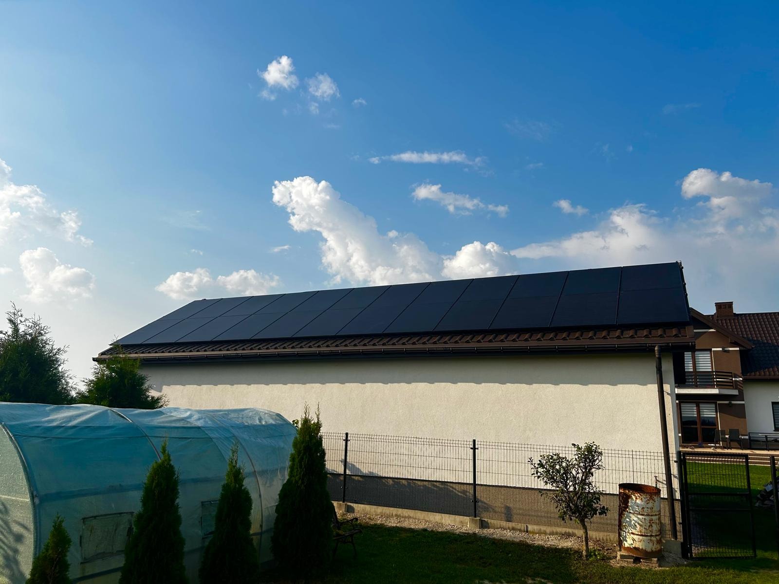 Czarny panel słoneczny na dachu jasnego budynku z brązowymi rynnami, widok z dołu na tle błękitnego nieba z białymi obłokami, obok szklarni i metalowego ogrodzenia.