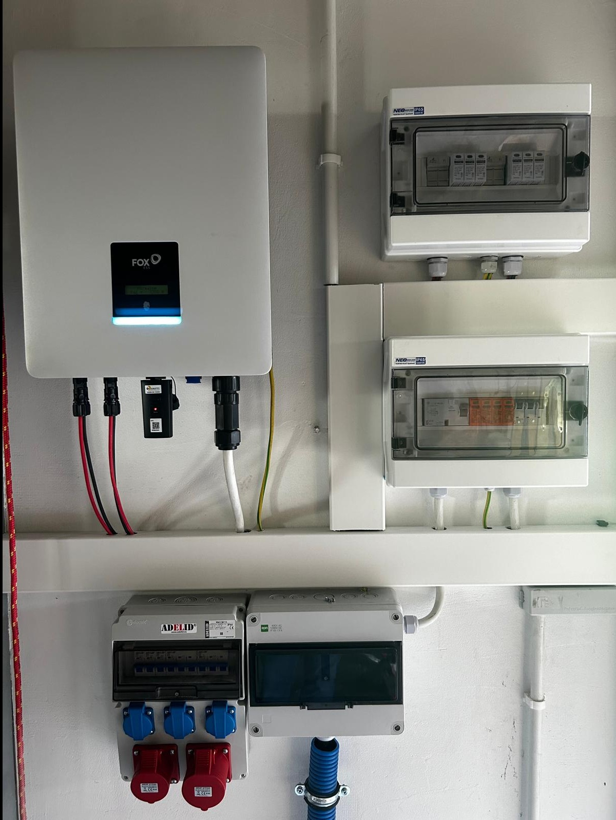 Biały falownik marki FOX z podłączonymi kablami, zamontowany na ścianie obok rozdzielnic elektrycznych z bezpiecznikami i gniazdami siłowymi.