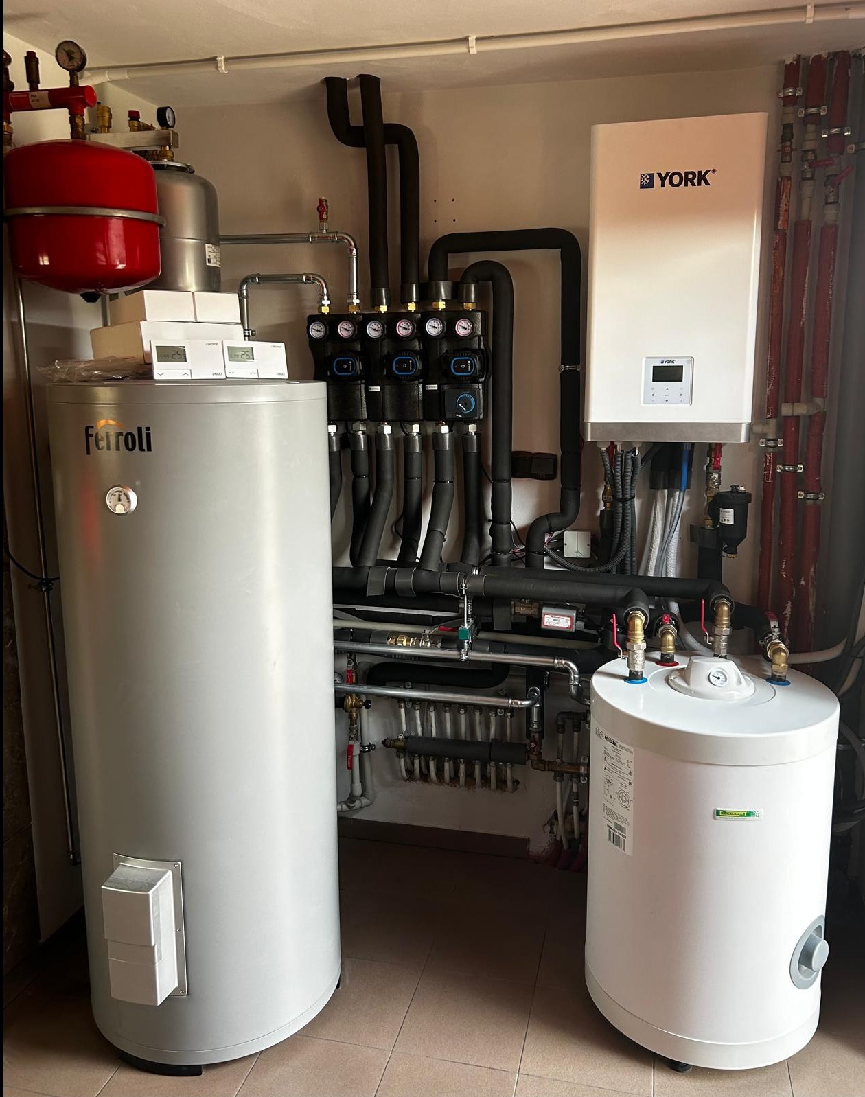 Kompletna instalacja grzewcza w kotłowni z zbiornikami Ferroli i York, rozdzielaczem z manometrami oraz licznymi rurami i zaworami.