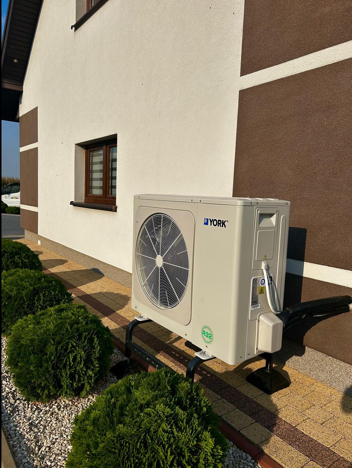 Zewnętrzna jednostka klimatyzacji marki York zamontowana na metalowym stelażu przy ścianie domu z brązowymi detalami, obok zadbane krzewy.