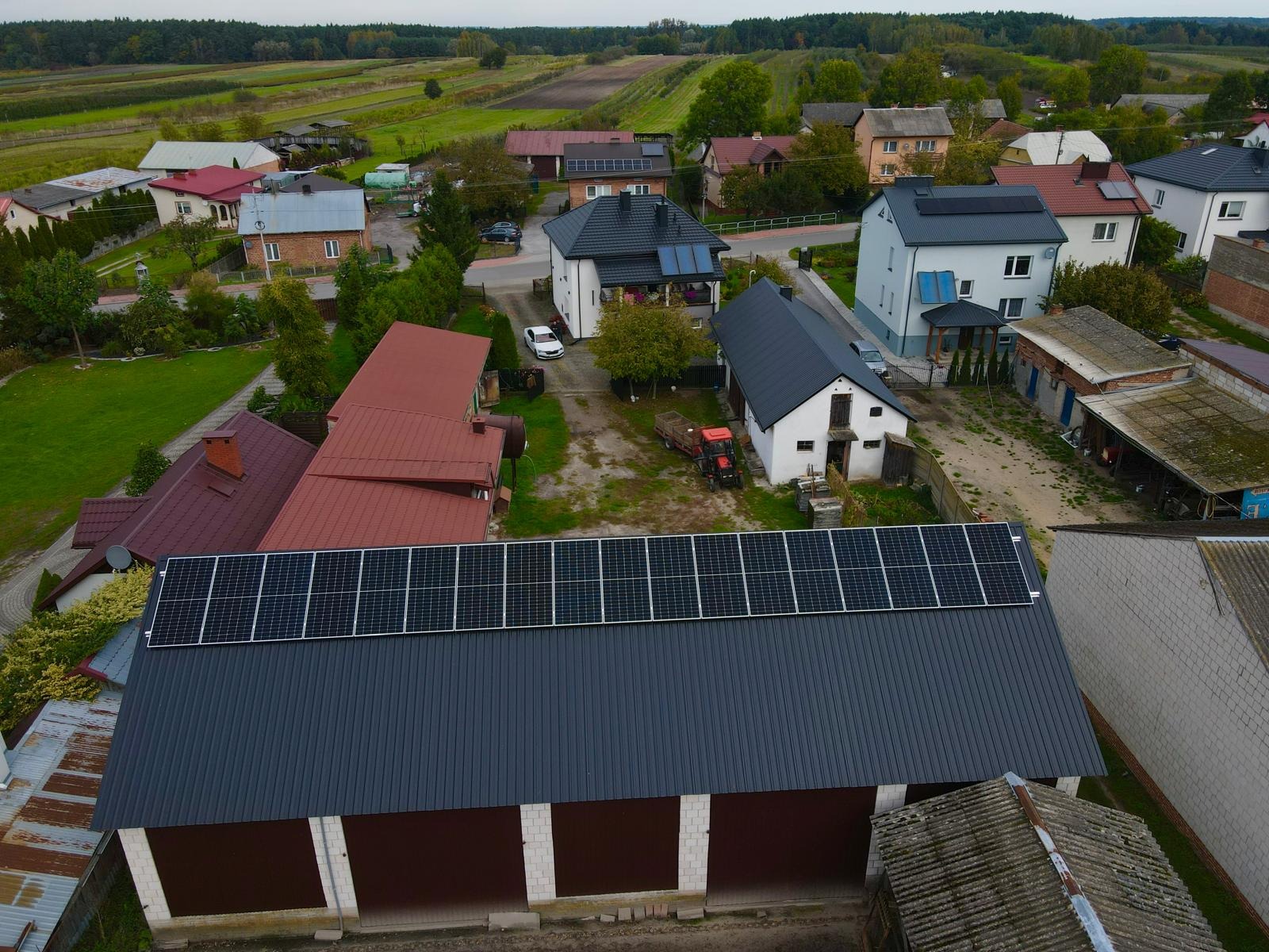 Widok z góry na wiejską zabudowę z panelami słonecznymi na dachu budynku gospodarczego i domu, w tle pola uprawne i las.