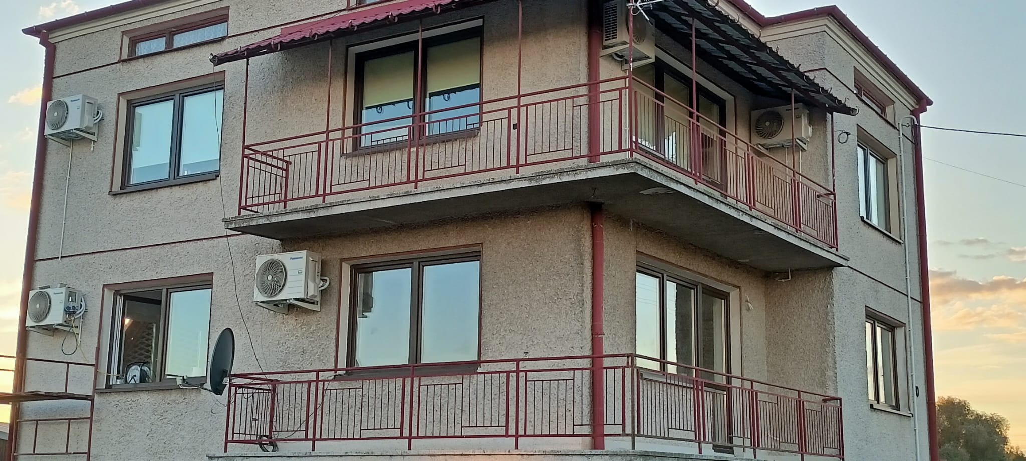 Dwupiętrowy budynek z oknami w ciemnych ramach, balkonami z czerwoną balustradą i zamontowanymi klimatyzatorami na zewnątrz.