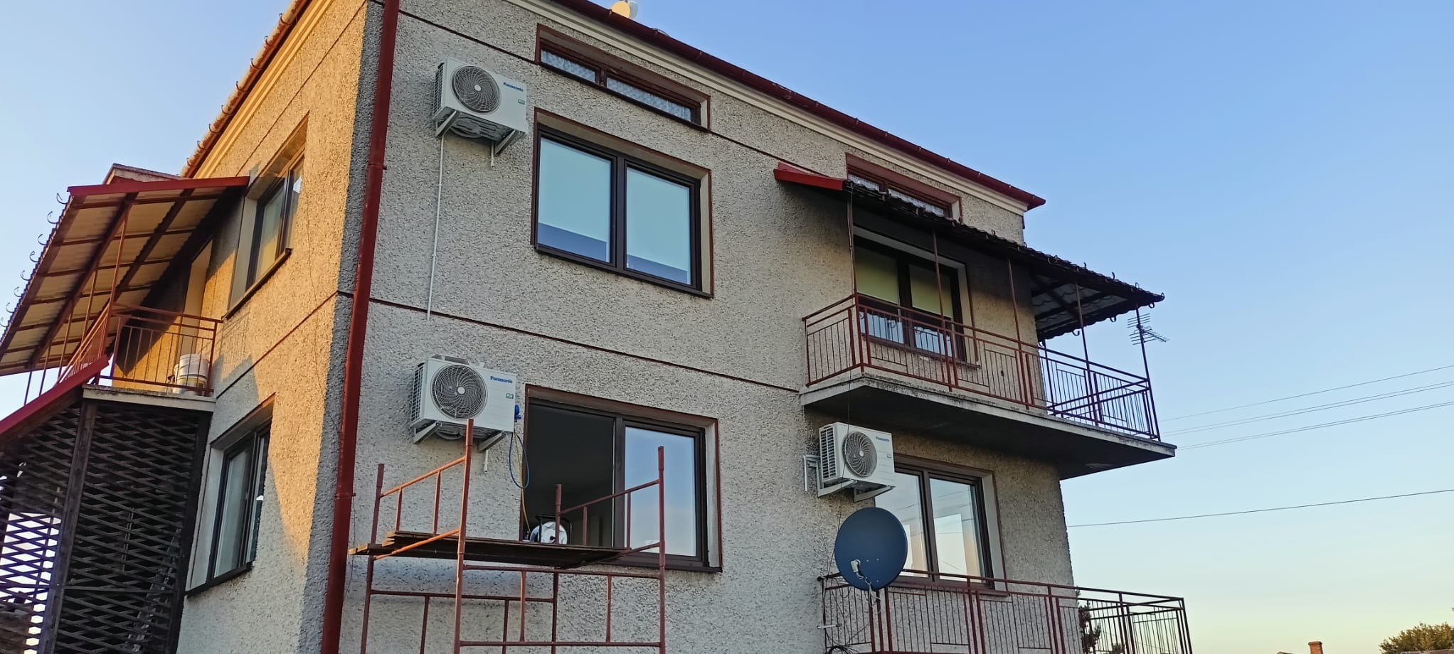 Elewacja domu z zainstalowanymi klimatyzatorami Panasonic, widoczne okna z brązowymi ramami, balkon z metalową balustradą i rusztowanie przy ścianie.