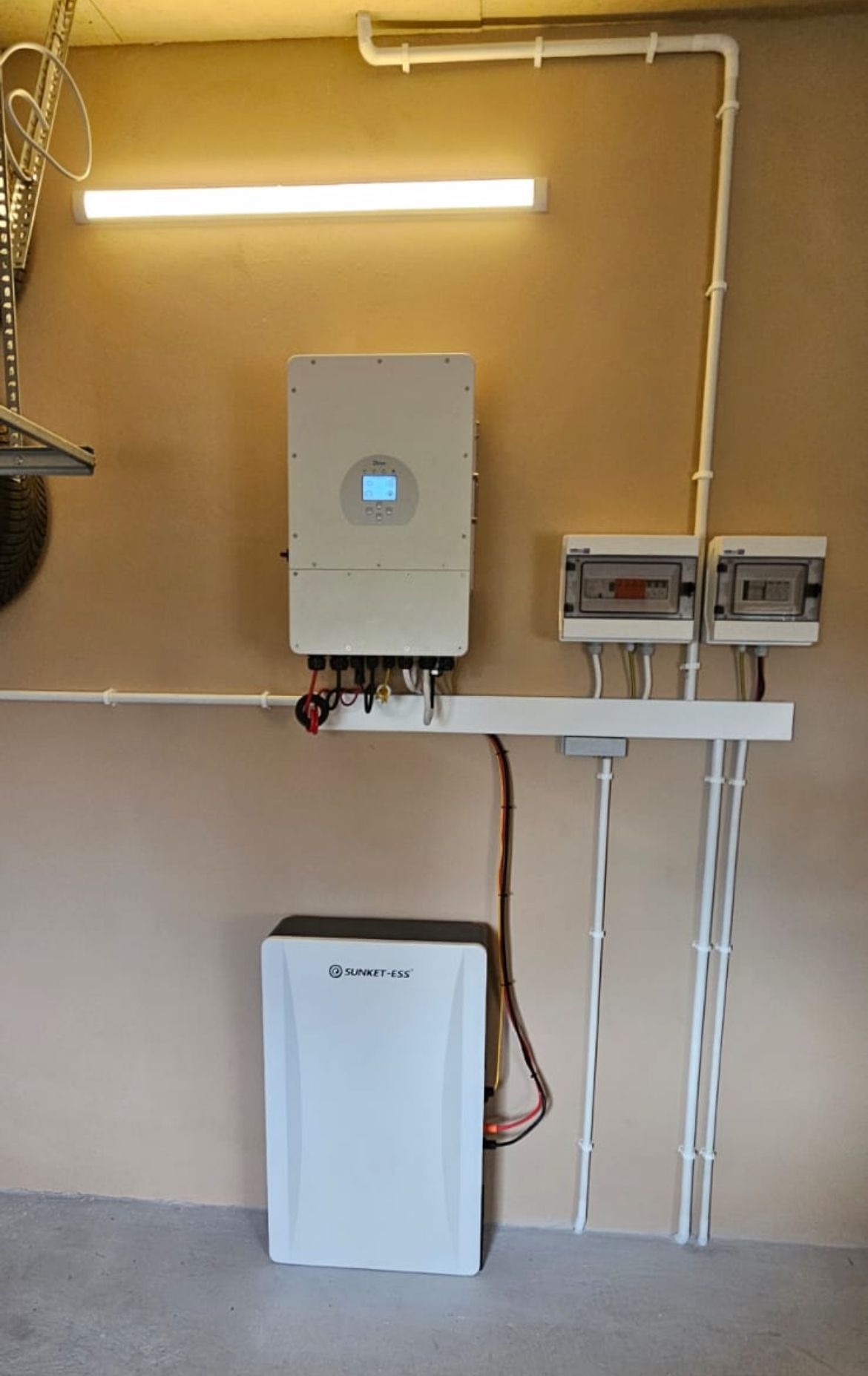 Montaż instalacji fotowoltaicznej w garażu: inwerter, akumulator i skrzynki bezpiecznikowe na beżowej ścianie, widoczne białe rury instalacyjne.