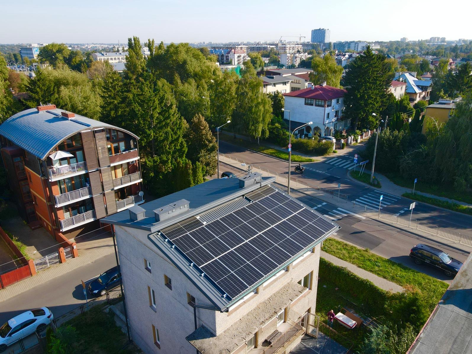 Widok z góry na budynek z zainstalowanymi panelami słonecznymi na dachu, w tle zabudowa miejska i zieleń. Na pierwszym planie narożny budynek z teksturalną elewacją. W tle widoczny ruch uliczny.