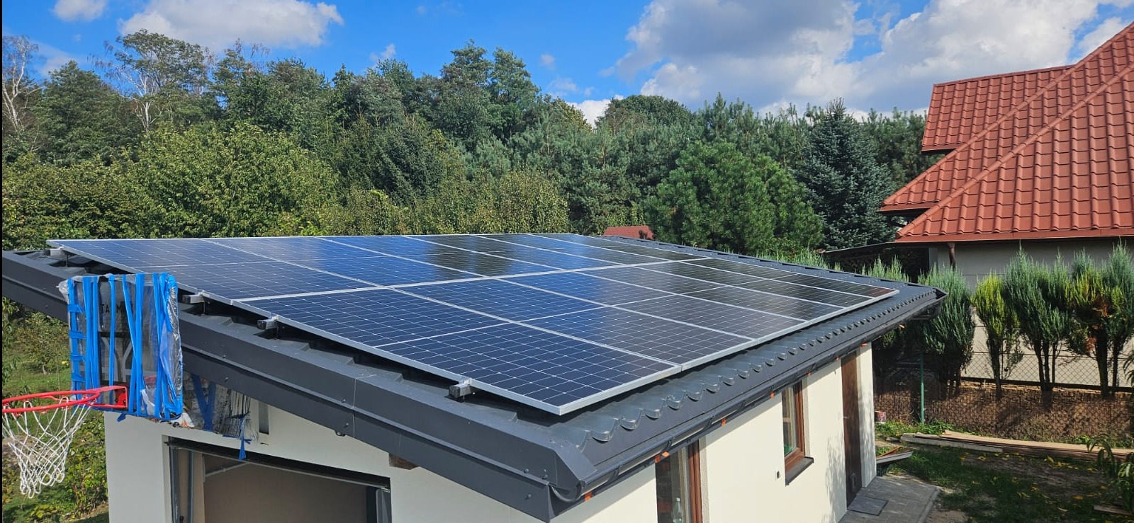 Panele słoneczne na dachu budynku z widocznym koszem do koszykówki zabezpieczonym folią, na tle zieleni i błękitnego nieba.