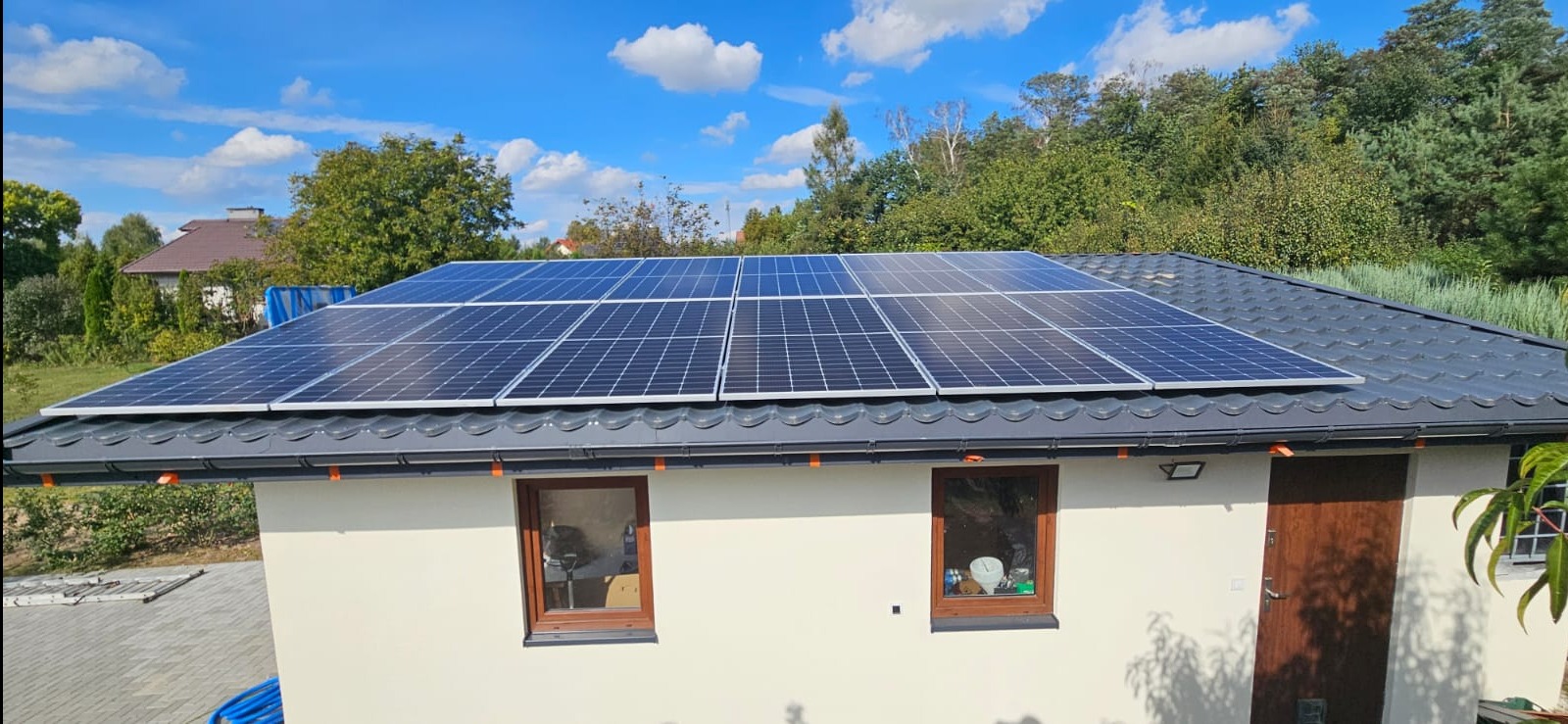 Panele fotowoltaiczne zamontowane na dachu budynku z szarą dachówką, widoczne brązowe okna i drzwi, błękitne niebo z chmurami w tle.