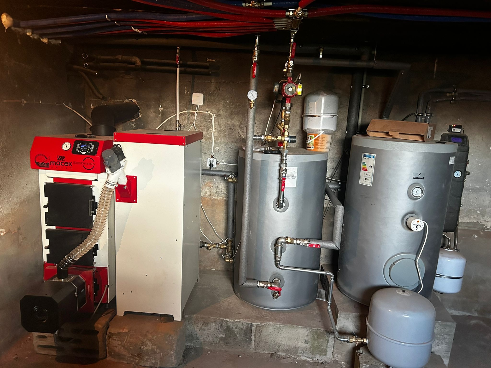 Instalacja centralnego ogrzewania z kotłem na paliwo stałe i dwoma zbiornikami akumulacyjnymi w piwnicy z widocznymi rurami instalacji grzewczej pod sufitem.
