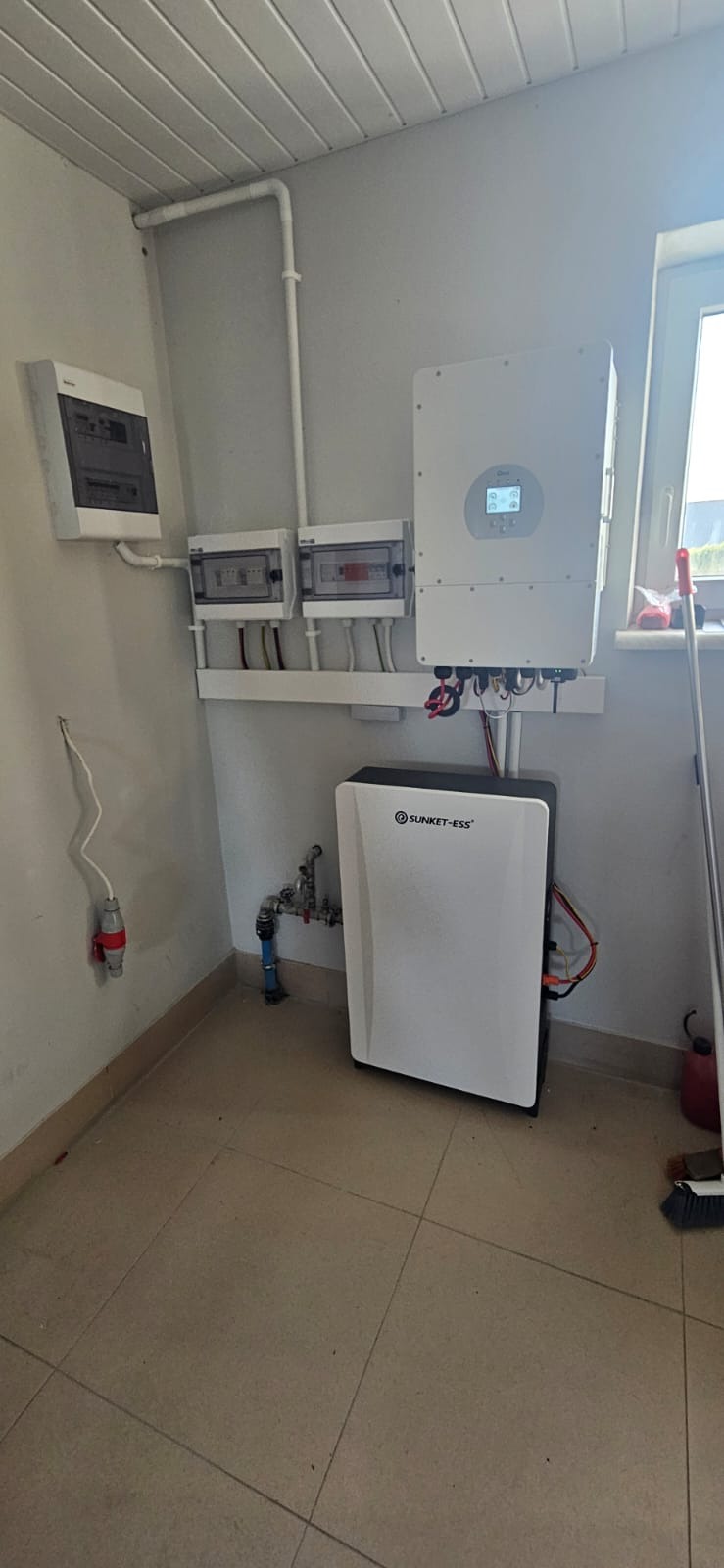 Wnętrze pomieszczenia z zainstalowanym inwerterem i akumulatorem energii marki SUNKET-ESS, widoczne również skrzynki elektryczne i fragment instalacji wodnej.