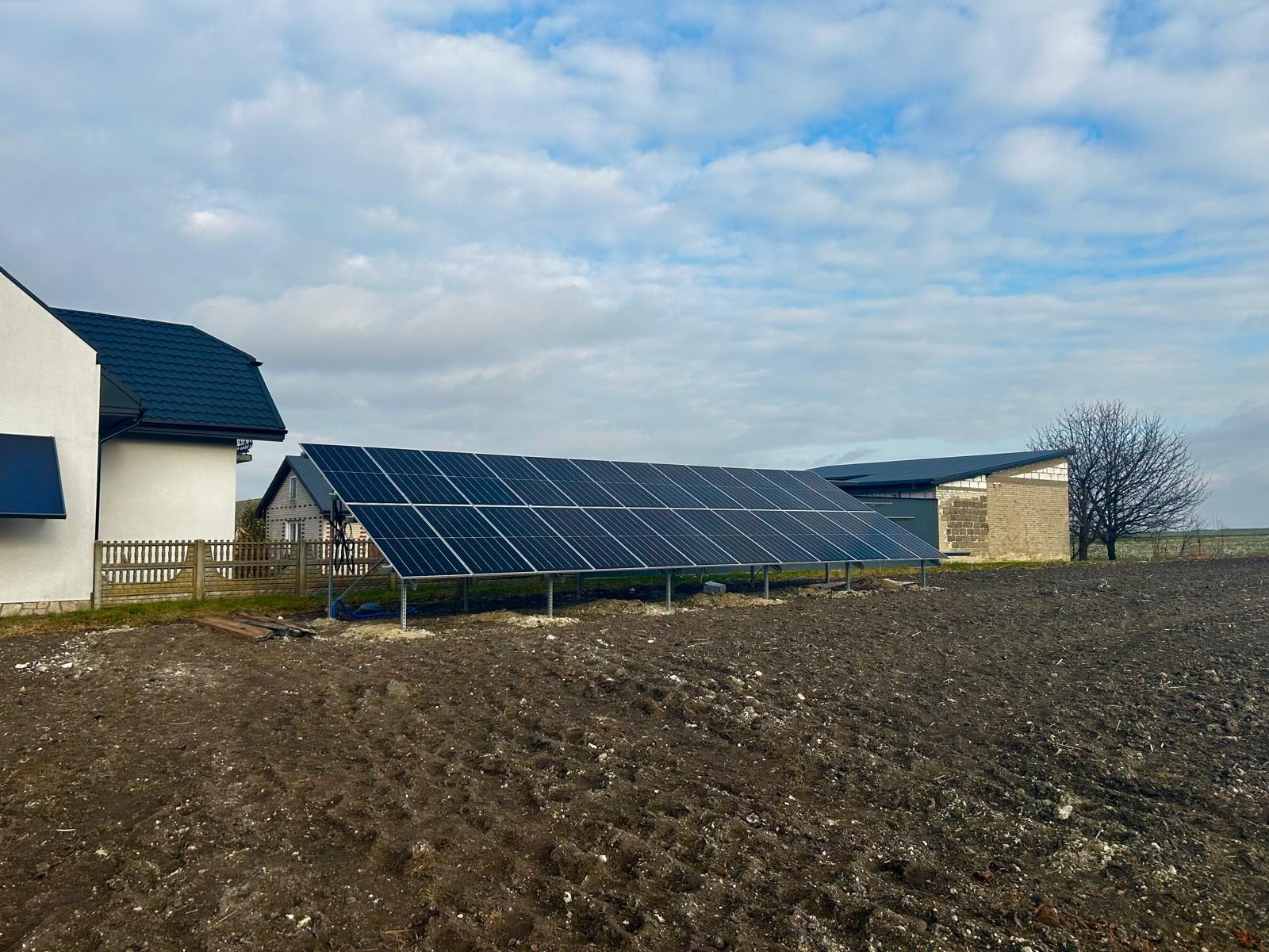 Instalacja fotowoltaiczna gruntowa z czarnymi panelami na metalowym stelażu, w tle budynki mieszkalne i pole orne pod pochmurnym niebem.