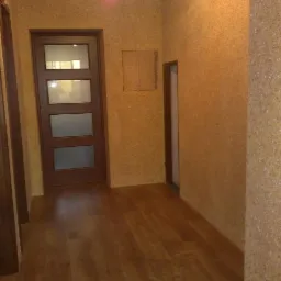 Korytarz z tapetą natryskową w odcieniach beżu i brązu, drewniana podłoga, drzwi z przeszkleniami i widocznym fragmentem ościeżnicy.