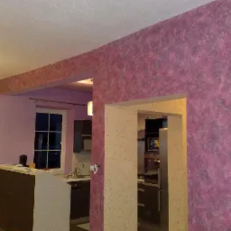 Fragment wnętrza po malowaniu dekoracyjnym, ściana w odcieniach fioletu z efektem struktury, widok na kuchnię przez otwór w ścianie.